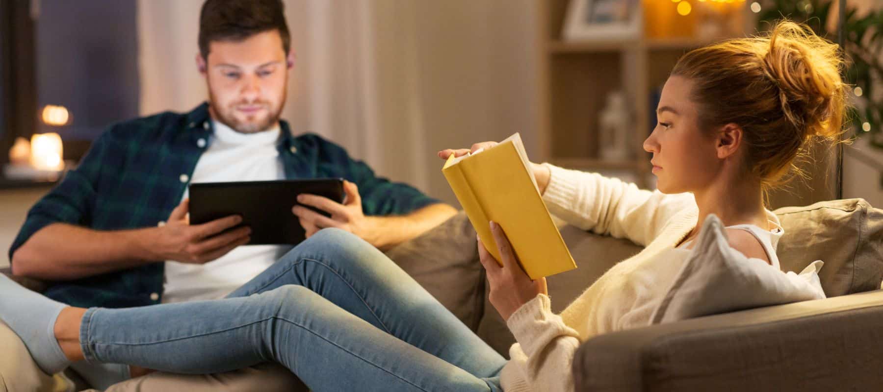 couple comfortable on their couch reading a book and looking at their tablet, comfortable because of heating services from hill and company